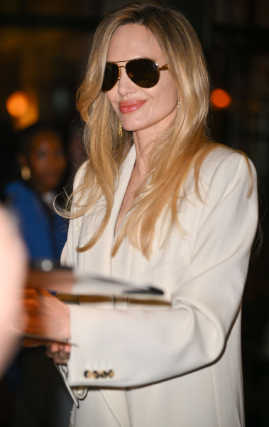 Angelina Jolie signs autographs in a white coat. She has blonde hair and wears sunglasses.